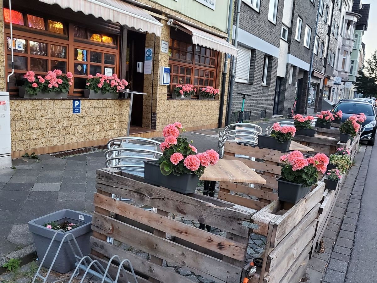 Sitzplätze vor der Gaststätte, eingefasst mit Holzkästen voller rosa Geranien, dahinter die Blumenkästen an den Fenstern.