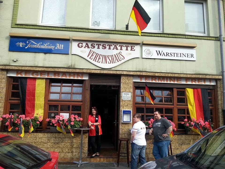 Die Gaststätte von außen: das Schild über der Tür, daneben die Schilder von Frankenheim Alt und Warsteiner, Geranien in den Fensterkästen.
